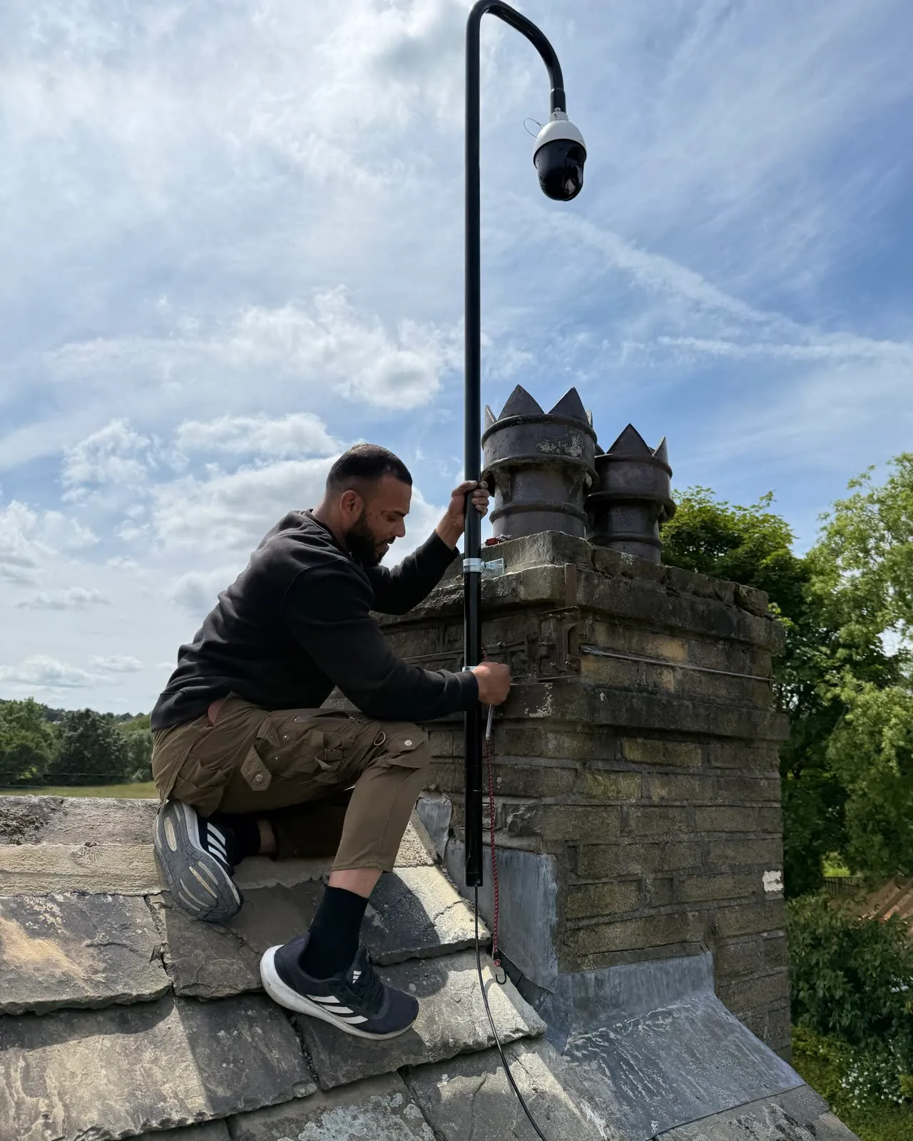 Mitu sat on the roof beside a stone chimney stack, fixing the pole for a Hikvision PTZ camera